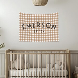 May include: A tan and white checkered wall tapestry with the name "EMERSON" above "SUTTON" in black lettering. The tapestry hangs above a wooden crib with white bedding and pillows. A plant and a woven basket are in the foreground.