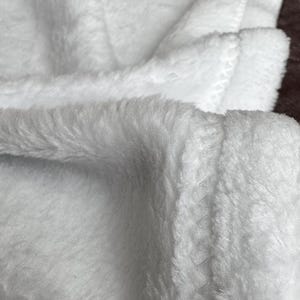 May include: Close-up of a soft, white blanket with a plush, fuzzy texture. The blanket is folded, revealing multiple layers and stitched edges. The fabric appears warm and inviting, perfect for cozy comfort.