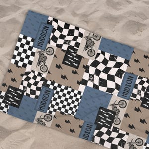 May include: Beach towel with a patchwork design featuring black and white checkered patterns, motorcycles, and lightning bolts. The name "HUDSON" is printed on blue rectangular patches. The towel is on a sandy surface.