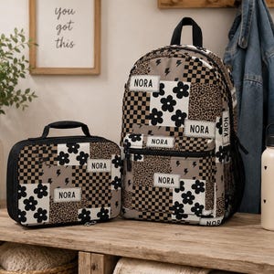 May include: A backpack and a matching lunch bag featuring a patchwork design of black and white flowers, brown and black checkerboard patterns, leopard print, and lightning bolts. Each item has a white rectangular patch with the name NORA.