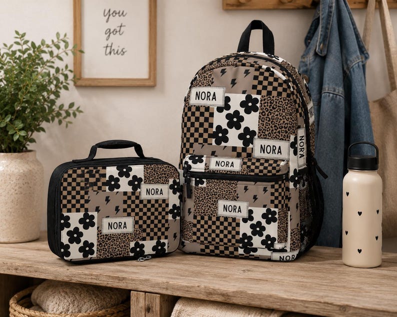 May include: A backpack and a matching lunch bag featuring a patchwork design of black and white flowers, brown and black checkerboard patterns, leopard print, and lightning bolts. Each item has a white rectangular patch with the name NORA.