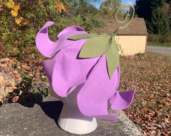 Può includere: Un cappello di feltro viola a forma di fiore con uno stelo e una foglia di feltro verde. Il cappello è su una testa di manichino bianca.