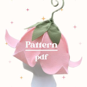 May include: A pink felt flower hat with green leaves and a curled stem. The hat is on a white mannequin head. Text on the hat reads "Pattern pdf". The background is light with star accents.