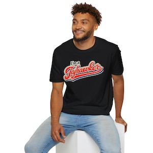 May include: A black short-sleeved t-shirt featuring the text "I'm a Jayhawker" printed across the chest. The word "Jayhawker" is written in a large, stylized red script font with a white and dark blue outline, while "I'm a" appears above in blue.