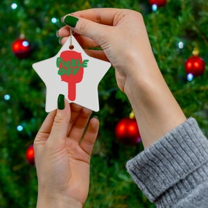 May include: A white star-shaped Christmas ornament with the words "Pickle Off" written in green and red letters. The ornament is being held by a hand with green nail polish.