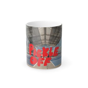 May include: White ceramic mug with a wraparound image of a pickleball court under a large, arched roof. Bold red text on the mug reads "PICKLE OFF". The mug is a standard size for hot beverages.