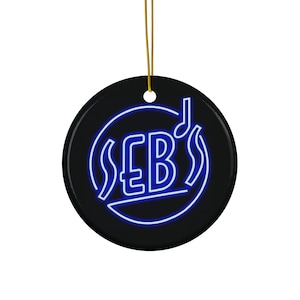 May include: A black, round ornament with a gold string for hanging. The ornament features the letters "SEB'S" in a blue neon-style font, encircled by a blue outline. A musical note is above the letters.