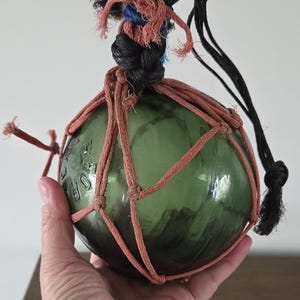 May include: A spherical green glass fishing float encased in a decorative knotted net made of salmon-coloured cord. The top features a thick black braided rope handle with frayed ends. Embossed text is visible on the side of the glass surface.