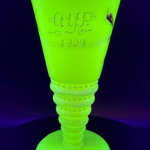 Puede incluir: Un jarrón de vidrio verde neón vibrante con una parte superior acampanada y una base redonda. El jarrón presenta un tallo texturizado y la inscripción "A.U.P. 1909". El borde tiene un borde festoneado, y el jarrón brilla intensamente sobre un fondo oscuro.