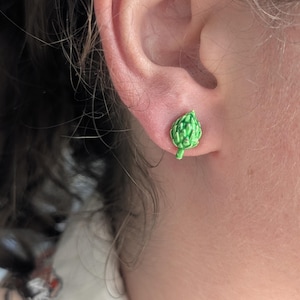 Peut inclure: Une petite boucle d'oreille à clou en forme d'artichaut vert. La boucle d'oreille présente une surface texturée et détaillée avec des nuances variées d'émail vert clair et foncé, reproduisant les feuilles superposées et la tige du légume.
