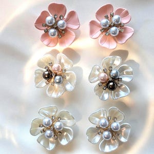 May include: Six flower-shaped earrings with pearl and rhinestone detailing. Two pairs are pale pink, and the other two pairs are white with a pearlescent sheen. Each earring features a cluster of pearls in the centre.