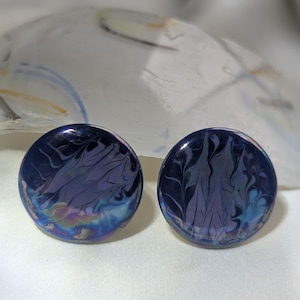 Peut inclure: Une paire de boucles d'oreilles à clous circulaires présentant une surface bleu foncé brillante avec des motifs marbrés tourbillonnants dans des tons plus clairs de bleu et de violet, créant un design fluide et abstrait sur les faces lisses.