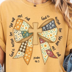 Può includere: T-shirt giallo senape con un grande design a fiocco e una croce. Il fiocco è realizzato con motivi patchwork con disegni floreali e a quadri. Il testo include "God is within her She will not fall" e "salmo 46:5".