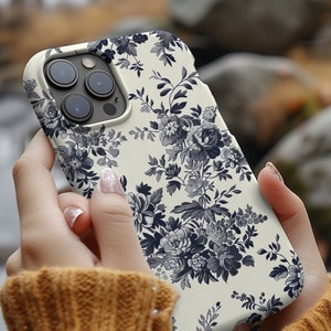 May include: A white iPhone case with a black and white floral pattern. The case has a raised camera bump and a cutout for the charging port.