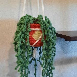 Puede incluir: Una maceta colgante de ganchillo hecha a mano con una maceta roja y amarilla, adornada con vides de ganchillo verdes y colgantes. La maceta está suspendida por cuerdas blancas, creando una pieza decorativa única para plantas de interior.