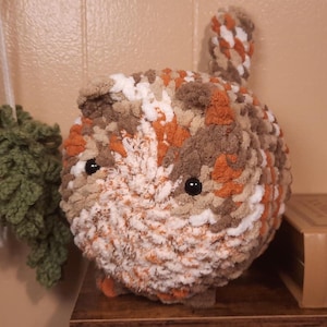 May include: A handmade, round crochet cat plushie in shades of brown, orange, and white. The plushie has black button eyes and a small, upright tail. The cat is sitting on a wooden surface.