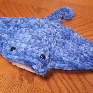 May include: A plush stingray toy crafted from soft, textured blue yarn. The creature features a wide, flat body with two small, round black plastic eyes positioned on the top and a white underside visible near the mouth area, ending in a curved tail.