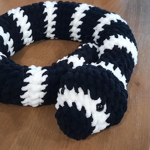May include: A plush snake toy crafted from soft, chunky yarn featuring a repeating pattern of thick black and white stripes. The stuffed animal has a coiled body and two small, round, black plastic eyes positioned on the front of its head.