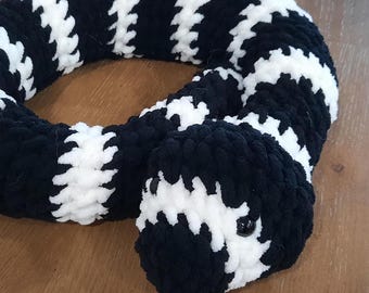 May include: A plush snake toy crafted from soft, chunky yarn featuring a repeating pattern of thick black and white stripes. The stuffed animal has a coiled body and two small, round, black plastic eyes positioned on the front of its head.