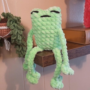 May include: A light green crocheted frog with long legs sits on a wooden shelf. A hanging crocheted plant and a red crocheted figure are also on the shelf. The frog has closed eyes and a relaxed pose.