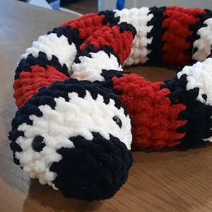 May include: A plush snake toy crafted from thick, soft yarn featuring a repeating pattern of bold red, stark white, and deep black segments. The stuffed creature has a rounded head with two small, smooth, black plastic eyes set into the white yarn.