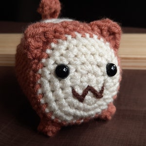 May include: A small crocheted stuffed toy with brown and white fur, black eyes, and a brown stitched smile.