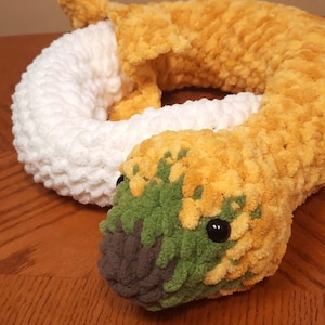 May include: A coiled plush snake toy crafted from soft, chunky yarn. The body features alternating sections of bright yellow and white. The head is yellow with patches of dark green and a dark gray snout, accented by small, round, black plastic eyes.