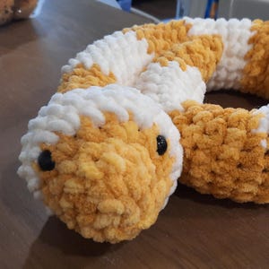 May include: A handmade, plush snake toy in shades of yellow, white, and black. The snake's body is a soft, textured material with a pattern of yellow and white sections. It has black button eyes.