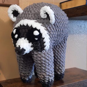 May include: A handmade crochet raccoon plush toy featuring a grey body, a white face mask, black eye patches, and white-tipped ears. The stuffed animal is crafted from thick, textured yarn and stands on four legs with dark grey paws at the base.