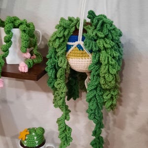 May include: A crocheted hanging planter with green leaves and a colorful patterned pot. The planter is hanging from a white string and is displayed on a shelf with two other crocheted plants.