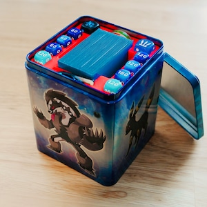 May include: A blue metal box with a hinged lid, filled with game dice and cards. The box features a galaxy design and cartoon character illustrations. The dice are blue and teal, with numbers indicating game values.