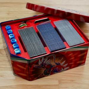 May include: A red and white metal box with a dragon design. The box is open and contains three stacks of cards and a set of blue dice with numbers 20, 40, 60, 80, 140, and 180.