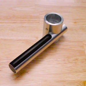 May include: A silver-colored metal object with a cylindrical top and a long, rectangular handle. The handle has a black, recessed channel running its length. The object is set against a light brown wooden surface.