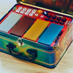 May include: A blue and gold metal tin with a Pokemon character on the side. The tin is open and contains a deck of cards, dice, and counters.