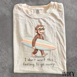 May include: Ivory-colored t-shirt featuring a cartoon monkey wearing sunglasses and carrying a surfboard with pastel stripes. The text "I don't want this feeling to go away" is printed below the graphic. The shirt is folded.