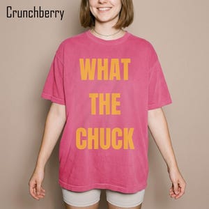 May include: A short-sleeved, crew-neck t-shirt in a vibrant pink color. The front of the shirt features the text "WHAT THE CHUCK" printed in large, bold, yellow capital letters. The word "Crunchberry" is also visible in the top left corner.