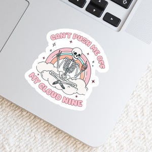 May include: A die-cut sticker featuring a skeleton meditating on a cloud with a rainbow and the text "Can't Push Me Off My Cloud Nine." The skeleton is holding a peace sign and a coffee cup. The sticker has a pastel color scheme.