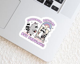 May include: A die-cut sticker featuring two illustrated raccoons, one standing outside and one reflected in a mirror, both holding drinks. The design includes a rainbow, clouds, and the text "EXTREMELY BIPOLAR BUT FABULOUS" in purple letters.