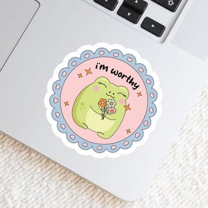 May include: A circular sticker featuring a light green frog holding a small bouquet of flowers. The background is light pink with orange star accents. The text "i'm worthy" is printed in black. The border is a scalloped blue frame with hearts.