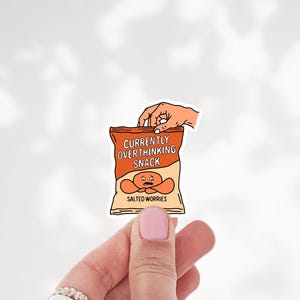 May include: A die-cut sticker featuring an orange snack bag with the text "CURRENTLY OVERTHINKING SNACK" and "SALTED WORRIES" printed on the front. The bag design includes a cartoon character with a worried expression and a hand reaching inside.
