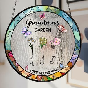 May include: Circular stained glass garden sign with the text "Grandma's Garden". The sign features butterflies, flowers, and the names Amelia, Olivia, and Daisy. The sign is bordered with a colourful mosaic design and hangs from a chain.