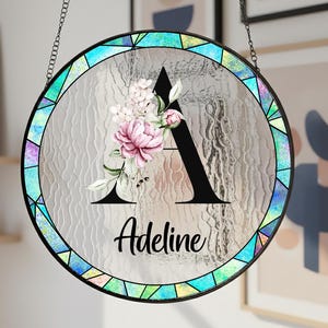 May include: Circular stained glass window with a black metal frame and chain. It displays a black letter "A" with a floral design and the name "Adeline". The border has a colourful geometric pattern. The window hangs indoors.