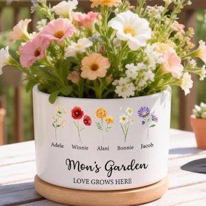May include: A white ceramic flower pot with a wooden base, filled with colourful flowers. The pot is decorated with floral illustrations and the text "Mom's Garden, Love Grows Here". The names Adele, Winnie, Alani, Bonnie, and Jacob are also printed on the pot.