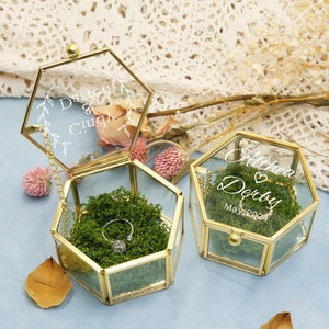 May include: Two hexagonal glass ring boxes with gold trim. The tops of the boxes are engraved with the names "Althea & Derby" and "D'Lishu & Cindy". The boxes are filled with green moss and a silver ring is in one of the boxes.