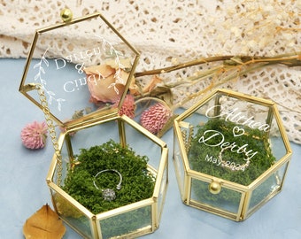 May include: Two hexagonal glass ring boxes with gold trim. The top of the boxes are engraved with the names "Althea & Derby" and "D'Lishu & Cindy". The boxes are filled with green moss and a silver ring is in one of the boxes.
