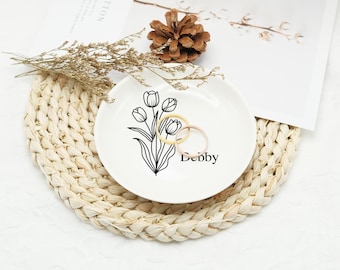 May include: A white ceramic ring dish with a black floral tulip design and the name "Debby" in black script. Two rings rest on the dish. The dish sits on a woven placemat, with a pinecone and dried flowers nearby.