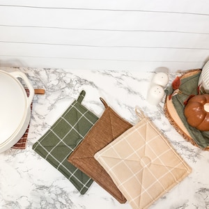 Op de afbeelding: Drie ovenwanten in verschillende kleuren en patronen. De ovenwanten zijn groen met witte ruitjes, bruin met witte ruitjes en beige met witte ruitjes. De ovenwanten liggen op een wit marmeren oppervlak.