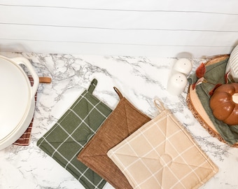 May include: Three pot holders in different colors and patterns. The pot holders are green with white checks, brown with white checks, and beige with white checks. The pot holders are on a white marble surface.