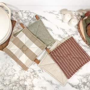 May include: Four different patterned kitchen pot holders with brown leather hanging loops. The pot holders are in a checkered pattern, a striped pattern, a solid green pattern, and a striped pattern with a brown background.