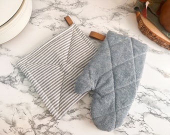 May include: Two kitchen pot holders, one is a striped fabric with a brown leather loop and the other is a gray quilted fabric with a brown leather loop.